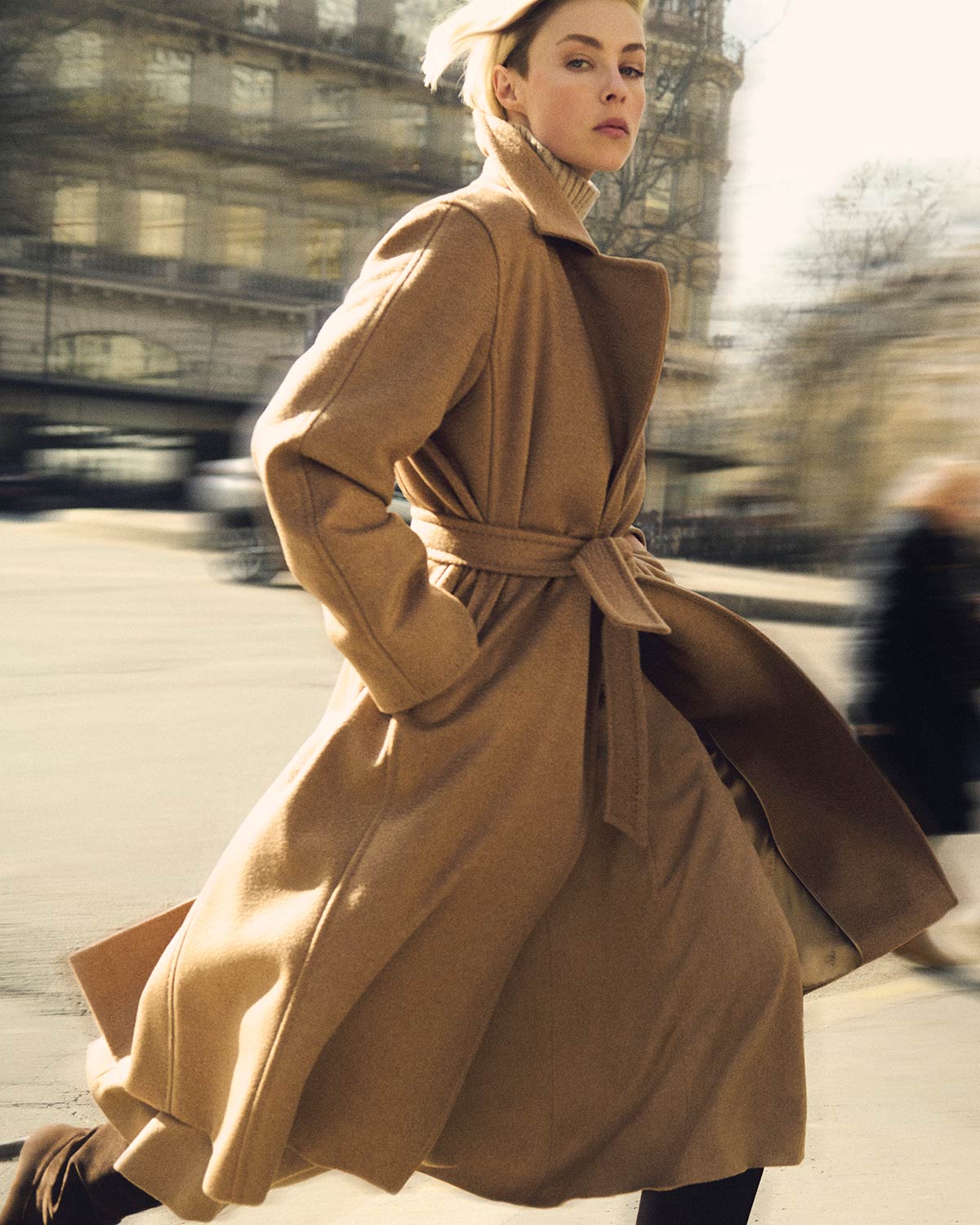 Mannequin portant le manteau iconique Manuela de Max Mara couleur chameau noué à la taille à l’allure élégante et dynamique en ville.