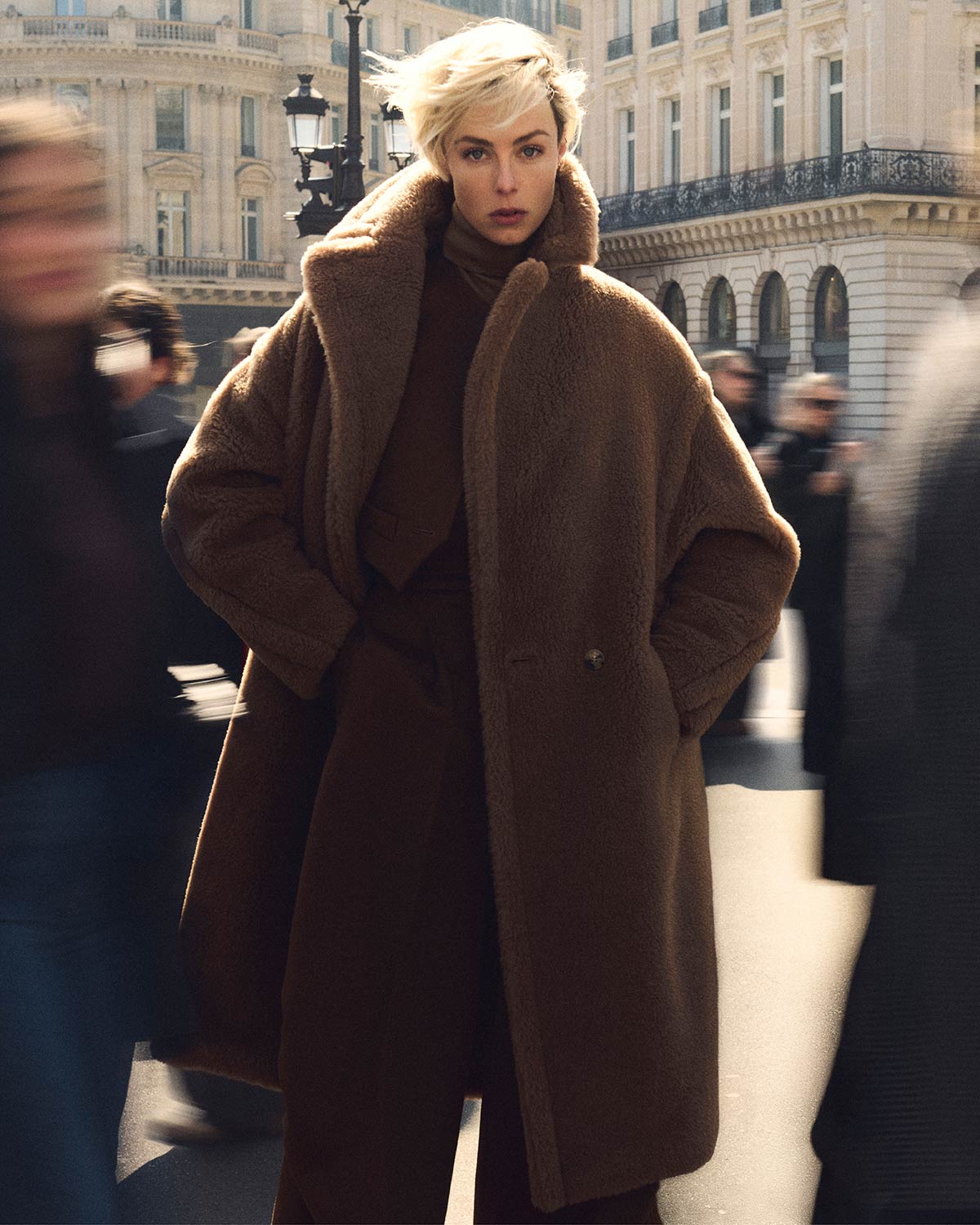 Mannequin portant le manteau iconique Teddy Bear long de Max Mara couleur chameau en milieu citadin bondé.